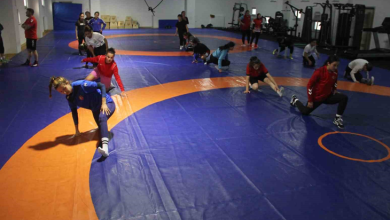 Görselin üst bölümünde, kapalı spor salonundaki mavi ve turuncu zeminli güreş minderi üzerinde antrenman yapan İşitme Engelli Kadın Güreş Milli Takımı sporcuları ve antrenörleri yer alıyor. Ön planda mavi ve kırmızı antrenman formaları giymiş kadın güreşçiler minderde teknik hareket ve taktik çalışması yaparken; orta alanda beyaz tişörtlü bir sporcu diğer takım arkadaşlarıyla ilgileniyor. Sol tarafta ise kırmızı tişörtlü ve siyah şortlu erkek antrenör kollarını kavuşturmuş vaziyette sporcuların çalışmasını dikkatle takip ediyor. Arka fonda mavi koruma minderleri, ahşap antrenman kasaları ve spor salonunun geniş iç mekânı görülüyor.