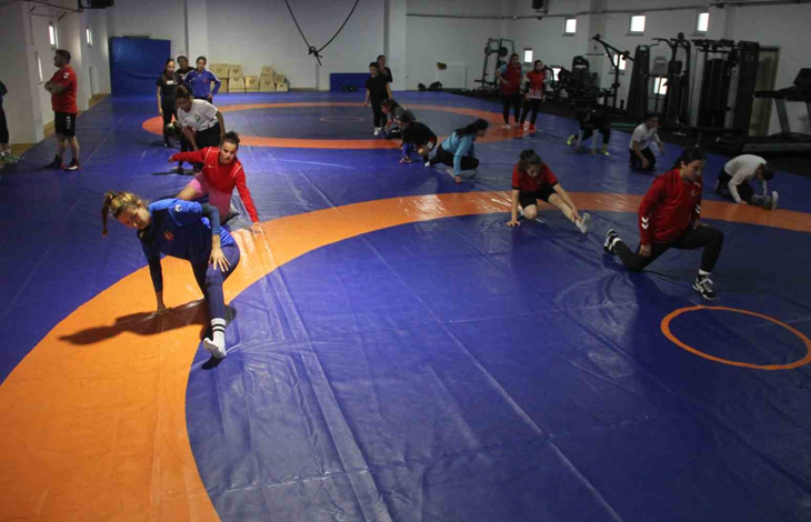 Görselin üst bölümünde, kapalı spor salonundaki mavi ve turuncu zeminli güreş minderi üzerinde antrenman yapan İşitme Engelli Kadın Güreş Milli Takımı sporcuları ve antrenörleri yer alıyor. Ön planda mavi ve kırmızı antrenman formaları giymiş kadın güreşçiler minderde teknik hareket ve taktik çalışması yaparken; orta alanda beyaz tişörtlü bir sporcu diğer takım arkadaşlarıyla ilgileniyor. Sol tarafta ise kırmızı tişörtlü ve siyah şortlu erkek antrenör kollarını kavuşturmuş vaziyette sporcuların çalışmasını dikkatle takip ediyor. Arka fonda mavi koruma minderleri, ahşap antrenman kasaları ve spor salonunun geniş iç mekânı görülüyor.