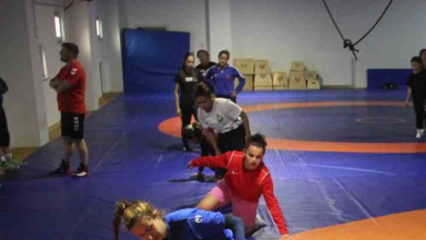 Görselin üst bölümünde, kapalı spor salonundaki mavi ve turuncu zeminli güreş minderi üzerinde antrenman yapan İşitme Engelli Kadın Güreş Milli Takımı sporcuları ve antrenörleri yer alıyor. Ön planda mavi ve kırmızı antrenman formaları giymiş kadın güreşçiler minderde teknik hareket ve taktik çalışması yaparken; orta alanda beyaz tişörtlü bir sporcu diğer takım arkadaşlarıyla ilgileniyor. Sol tarafta ise kırmızı tişörtlü ve siyah şortlu erkek antrenör kollarını kavuşturmuş vaziyette sporcuların çalışmasını dikkatle takip ediyor. Arka fonda mavi koruma minderleri, ahşap antrenman kasaları ve spor salonunun geniş iç mekânı görülüyor.