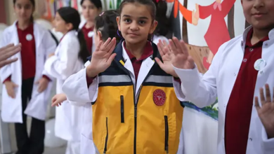 Görselin üst bölümünde, beyaz doktor önlükleri giymiş işitme engelli öğrenciler yer alıyor. Merkezde duran saçları toplu kız çocuğu, beyaz önlüğünün üzerine sarı ve gri renkli acil sağlık/ambulans görevlisi yeleği giymiş olarak iki elinin avuç içini ileriye doğru açarak işaret diliyle poz veriyor. Sağ ve sol tarafta beyaz önlük giymiş diğer kız ve erkek öğrenciler de ellerini kaldırarak işaret dilinde iletişime katılıyor. Arka planda renkli çizimlerin ve el ele tutuşan figürlerin bulunduğu bir duvar panosu görülüyor.