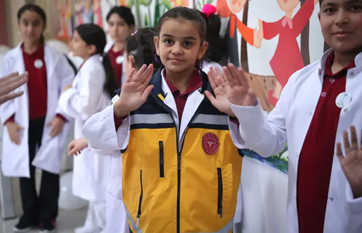 Görselin üst bölümünde, beyaz doktor önlükleri giymiş işitme engelli öğrenciler yer alıyor. Merkezde duran saçları toplu kız çocuğu, beyaz önlüğünün üzerine sarı ve gri renkli acil sağlık/ambulans görevlisi yeleği giymiş olarak iki elinin avuç içini ileriye doğru açarak işaret diliyle poz veriyor. Sağ ve sol tarafta beyaz önlük giymiş diğer kız ve erkek öğrenciler de ellerini kaldırarak işaret dilinde iletişime katılıyor. Arka planda renkli çizimlerin ve el ele tutuşan figürlerin bulunduğu bir duvar panosu görülüyor.