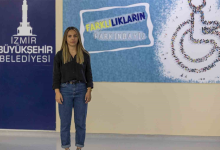 Görselin üst bölümünde, kapalı bir alanda, üzerinde "İZMİR BÜYÜKŞEHİR BELEDİYESİ" yazısı ve "FARKLILIKLARIN FARKINDAYIZ" afişi ile tekerlekli sandalye simgesi bulunan bir duvarın önünde ayakta durarak doğrudan kameraya bakan işitme engelli Sahra Nur Akyıl yer almaktadır. Siyah uzun kollu gömlek ve mavi kot pantolon giymiş olan Akyıl, hafifçe gülümseyerek poz vermektedir.