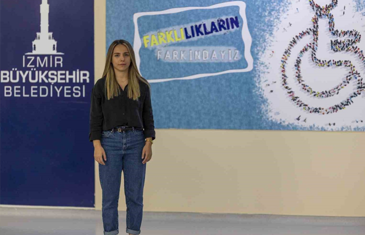 Görselin üst bölümünde, kapalı bir alanda, üzerinde "İZMİR BÜYÜKŞEHİR BELEDİYESİ" yazısı ve "FARKLILIKLARIN FARKINDAYIZ" afişi ile tekerlekli sandalye simgesi bulunan bir duvarın önünde ayakta durarak doğrudan kameraya bakan işitme engelli Sahra Nur Akyıl yer almaktadır. Siyah uzun kollu gömlek ve mavi kot pantolon giymiş olan Akyıl, hafifçe gülümseyerek poz vermektedir.