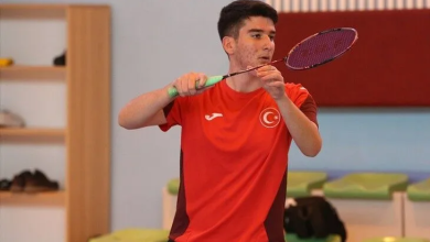 Görselin üst bölümünde, kapalı bir spor salonunda kırmızı-bordolu millî takım forması giymiş genç bir erkek badminton sporcusu elinde raketle vuruş pozisyonu almış halde yer almaktadır. Odaklanmış bir ifadeyle ileriye bakan sporcunun arkasında salon oturma alanları ve ahşap dolaplar görülmektedir.