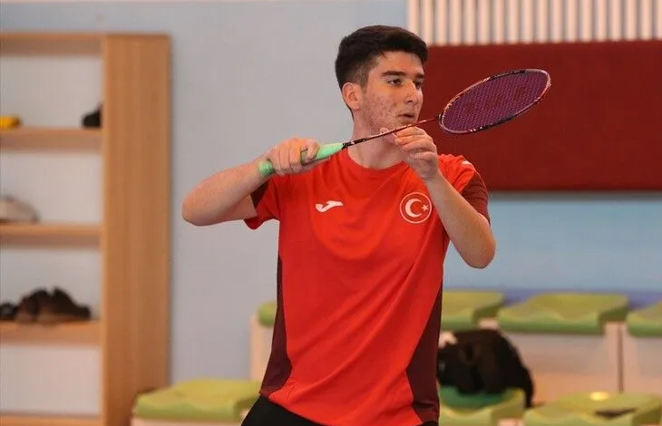 Görselin üst bölümünde, kapalı bir spor salonunda kırmızı-bordolu millî takım forması giymiş genç bir erkek badminton sporcusu elinde raketle vuruş pozisyonu almış halde yer almaktadır. Odaklanmış bir ifadeyle ileriye bakan sporcunun arkasında salon oturma alanları ve ahşap dolaplar görülmektedir.