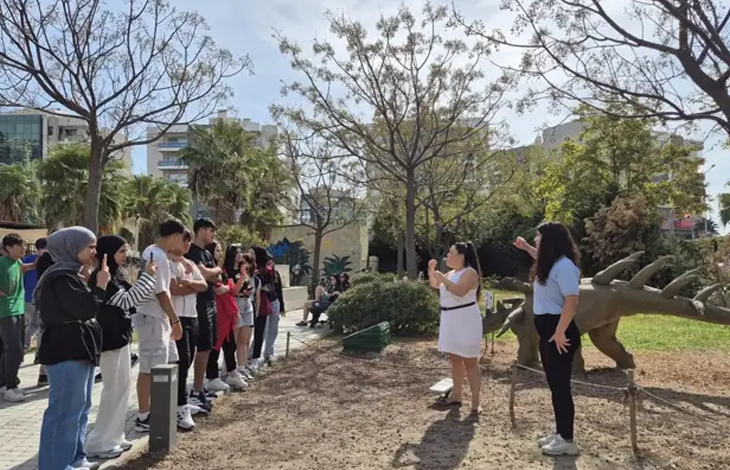 Görselin üst bölümünde, bir parkta toplanmış ve bir sunumu izleyen işitme engelli gençlerden oluşan bir grup yer almaktadır. Gruptaki katılımcılar pür dikkat dinlerken, iki işaret dili tercümanı, sağ tarafta sergilenen dinozor maketinin hemen önünde durarak, sunumdaki bilgileri işaret diliyle gruba aktarmaktadır. Arka planda ise parkın yeşil dokusu, ağaçlar ve şehir binaları görülmektedir.