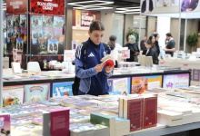 Görselin üst bölümünde, kalabalık ve geniş bir kitap fuarı alanında stantların arasında durarak elindeki kitabı inceleyen genç bir kadın kitapsever yer alıyor. Sırt çantası takmış ve kolları beyaz çizgili lacivert-mavi spor bir ceket giymiş olan saçları toplu genç kadın, önündeki geniş kitap masasının başında durarak iki eliyle açtığı kitabı dikkatle okuyor. Ön planda masaların üzerine üst üste dizilmiş çok sayıda ciltli ve renkli kitap yer alırken; arka planda yayınevlerinin kitap stantları, fuar afişleri ve fuarı gezen diğer ziyaretçiler görülüyor.