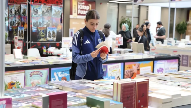 Görselin üst bölümünde, kalabalık ve geniş bir kitap fuarı alanında stantların arasında durarak elindeki kitabı inceleyen genç bir kadın kitapsever yer alıyor. Sırt çantası takmış ve kolları beyaz çizgili lacivert-mavi spor bir ceket giymiş olan saçları toplu genç kadın, önündeki geniş kitap masasının başında durarak iki eliyle açtığı kitabı dikkatle okuyor. Ön planda masaların üzerine üst üste dizilmiş çok sayıda ciltli ve renkli kitap yer alırken; arka planda yayınevlerinin kitap stantları, fuar afişleri ve fuarı gezen diğer ziyaretçiler görülüyor.