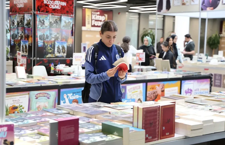Görselin üst bölümünde, kalabalık ve geniş bir kitap fuarı alanında stantların arasında durarak elindeki kitabı inceleyen genç bir kadın kitapsever yer alıyor. Sırt çantası takmış ve kolları beyaz çizgili lacivert-mavi spor bir ceket giymiş olan saçları toplu genç kadın, önündeki geniş kitap masasının başında durarak iki eliyle açtığı kitabı dikkatle okuyor. Ön planda masaların üzerine üst üste dizilmiş çok sayıda ciltli ve renkli kitap yer alırken; arka planda yayınevlerinin kitap stantları, fuar afişleri ve fuarı gezen diğer ziyaretçiler görülüyor.