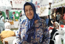 Görselin üst bölümünde, el emeği ürünlerin sergilendiği bir kermes veya pazar yerinde tezgâhının önünde durarak hafifçe gülümseyen engelli pazarcı Durdu Bayrak yer alıyor. Mavi tonlarında desenli bir elbise giyen ve lacivert başörtüsü takmış olan kadının başörtüsünün yakasında okuma gözlüğü asılı duruyor. Çevresinde ve başının üzerinde el işi danteller, işlemeli örtüler ve örgüler sergilenirken; arka planda beyaz plastik sandalyeler, stantları gezen vatandaşlar ve pazar alanının kalabalığı görülüyor.