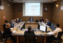 Görselin üst bölümünde, kurumsal bir disiplin veya encümen toplantı salonunda U şeklindeki ahşap masa etrafında toplanmış belediye yetkilileri ve komisyon üyeleri yer alıyor. Masanın başında takım elbiseli başkan otururken; her iki yanda kadın ve erkek kurul üyeleri, önlerindeki bilgisayar ekranları, evraklar, su şişeleri ve çay bardaklarıyla toplantıyı sürdürüyor. Arka plandaki duvara yansıtılan projeksiyon perdesinde Kocaeli Büyükşehir Belediyesi antetiyle birlikte konuya ilişkin resmi rapor ve tutanak maddeleri görülüyor; ahşap kaplama duvarlarda ise çerçeveli portreler ve duvar saatleri asılı duruyor.