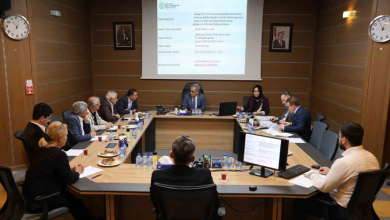 Görselin üst bölümünde, kurumsal bir disiplin veya encümen toplantı salonunda U şeklindeki ahşap masa etrafında toplanmış belediye yetkilileri ve komisyon üyeleri yer alıyor. Masanın başında takım elbiseli başkan otururken; her iki yanda kadın ve erkek kurul üyeleri, önlerindeki bilgisayar ekranları, evraklar, su şişeleri ve çay bardaklarıyla toplantıyı sürdürüyor. Arka plandaki duvara yansıtılan projeksiyon perdesinde Kocaeli Büyükşehir Belediyesi antetiyle birlikte konuya ilişkin resmi rapor ve tutanak maddeleri görülüyor; ahşap kaplama duvarlarda ise çerçeveli portreler ve duvar saatleri asılı duruyor.