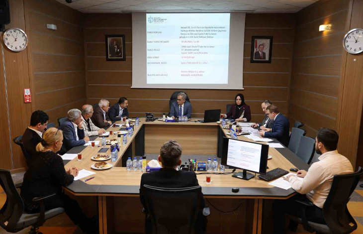 Görselin üst bölümünde, kurumsal bir disiplin veya encümen toplantı salonunda U şeklindeki ahşap masa etrafında toplanmış belediye yetkilileri ve komisyon üyeleri yer alıyor. Masanın başında takım elbiseli başkan otururken; her iki yanda kadın ve erkek kurul üyeleri, önlerindeki bilgisayar ekranları, evraklar, su şişeleri ve çay bardaklarıyla toplantıyı sürdürüyor. Arka plandaki duvara yansıtılan projeksiyon perdesinde Kocaeli Büyükşehir Belediyesi antetiyle birlikte konuya ilişkin resmi rapor ve tutanak maddeleri görülüyor; ahşap kaplama duvarlarda ise çerçeveli portreler ve duvar saatleri asılı duruyor.
