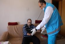 Görselde, mavi üniformalı bir sağlık görevlisi evde sağlık hizmeti kapsamında koltukta oturan hastaya fiziksel hareketler yaptırarak sağlık kontrolü gerçekleştirmektedir.