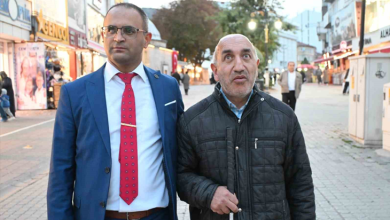 Görselin üst bölümünde, kalabalık bir cadde veya sokakta yan yana duran iki erkek yer almaktadır. Sol tarafta mavi takım elbise, beyaz gömlek ve kırmızı puantiyeli kravat giymiş, gözlüklü bir kişi; sağ tarafta ise siyah mont giyen ve elinde görme engelli bastonu tutan bir kişi bulunmaktadır. Arka planda dükkanlar, caddedeki yayalar ve sokak atmosferi görülmektedir.