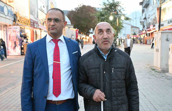 Görselin üst bölümünde, kalabalık bir cadde veya sokakta yan yana duran iki erkek yer almaktadır. Sol tarafta mavi takım elbise, beyaz gömlek ve kırmızı puantiyeli kravat giymiş, gözlüklü bir kişi; sağ tarafta ise siyah mont giyen ve elinde görme engelli bastonu tutan bir kişi bulunmaktadır. Arka planda dükkanlar, caddedeki yayalar ve sokak atmosferi görülmektedir.
