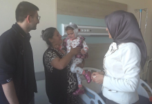 Görselde, bir hastane odasında yatağın yanında duran aile bireyleri ve başörtülü bir yetkili yer almaktadır. Annesinin kucağında tuttuğu, başı sargılı olan küçük bir çocuk ve sol tarafta duran bir erkek aile bireyi ile sağ taraftaki ziyaretçi yetkili, çocukla ilgilenirken görülmektedir.