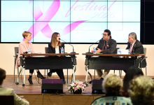 Bir konferans salonunda gerçekleştirilen panel oturumu görülmektedir. Sahnenin ön kısmında iki ayrı masada toplam dört panelist oturmaktadır. Her panelistin önünde mikrofon, su şişesi ve notlar bulunmaktadır. Masaların arasında pembe ve beyaz çiçeklerden oluşan bir aranjman yer almaktadır. Panelistlerin arkasında büyük bir dijital ekran bulunmaktadır. Ekranda pembe kurdele sembolü ve “festival 2026” yazısı görünmektedir; bu ifade etkinliğin meme sağlığı farkındalığıyla ilişkili olduğunu göstermektedir. Sahnenin önünde izleyiciler oturmakta ve paneli takip etmektedir. Ortam genel olarak profesyonel, düzenli ve bir sağlık farkındalığı etkinliğine uygun biçimde hazırlanmıştır.