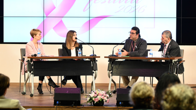Bir konferans salonunda gerçekleştirilen panel oturumu görülmektedir. Sahnenin ön kısmında iki ayrı masada toplam dört panelist oturmaktadır. Her panelistin önünde mikrofon, su şişesi ve notlar bulunmaktadır. Masaların arasında pembe ve beyaz çiçeklerden oluşan bir aranjman yer almaktadır. Panelistlerin arkasında büyük bir dijital ekran bulunmaktadır. Ekranda pembe kurdele sembolü ve “festival 2026” yazısı görünmektedir; bu ifade etkinliğin meme sağlığı farkındalığıyla ilişkili olduğunu göstermektedir. Sahnenin önünde izleyiciler oturmakta ve paneli takip etmektedir. Ortam genel olarak profesyonel, düzenli ve bir sağlık farkındalığı etkinliğine uygun biçimde hazırlanmıştır.