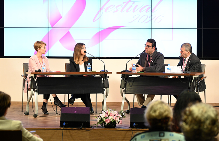 Bir konferans salonunda gerçekleştirilen panel oturumu görülmektedir. Sahnenin ön kısmında iki ayrı masada toplam dört panelist oturmaktadır. Her panelistin önünde mikrofon, su şişesi ve notlar bulunmaktadır. Masaların arasında pembe ve beyaz çiçeklerden oluşan bir aranjman yer almaktadır. Panelistlerin arkasında büyük bir dijital ekran bulunmaktadır. Ekranda pembe kurdele sembolü ve “festival 2026” yazısı görünmektedir; bu ifade etkinliğin meme sağlığı farkındalığıyla ilişkili olduğunu göstermektedir. Sahnenin önünde izleyiciler oturmakta ve paneli takip etmektedir. Ortam genel olarak profesyonel, düzenli ve bir sağlık farkındalığı etkinliğine uygun biçimde hazırlanmıştır.
