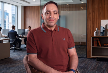 Mehmet Kızıltaş, ofis ortamında bir masada oturmuş şekilde kameraya doğru bakıyor. Üzerinde bordo bir polo tişört var. Arka planda cam bölmeli, modern dekorlu bir çalışma alanı ve bilgisayar başında çalışan iki kişi görülüyor. Ortam aydınlık, düzenli ve kurumsal bir ofis atmosferi taşıyor.
