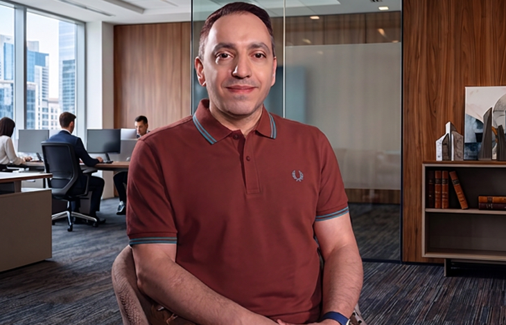 Mehmet Kızıltaş, ofis ortamında bir masada oturmuş şekilde kameraya doğru bakıyor. Üzerinde bordo bir polo tişört var. Arka planda cam bölmeli, modern dekorlu bir çalışma alanı ve bilgisayar başında çalışan iki kişi görülüyor. Ortam aydınlık, düzenli ve kurumsal bir ofis atmosferi taşıyor.