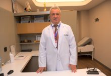 İzmir Özel Sağlık Hastanesi Ortopedi ve Travmatoloji Uzmanı Prof. Dr. Devrim Akseki, ofisinde ayakta durarak beyaz önlüğüyle poz veriyor.