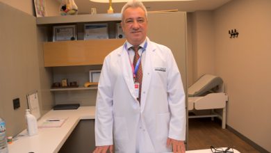 İzmir Özel Sağlık Hastanesi Ortopedi ve Travmatoloji Uzmanı Prof. Dr. Devrim Akseki, ofisinde ayakta durarak beyaz önlüğüyle poz veriyor.