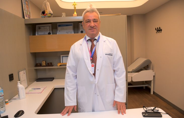 İzmir Özel Sağlık Hastanesi Ortopedi ve Travmatoloji Uzmanı Prof. Dr. Devrim Akseki, ofisinde ayakta durarak beyaz önlüğüyle poz veriyor.