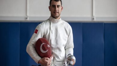 Türk bayrağı armalı beyaz eskrim kıyafeti giyen Hakan Akkaya, bir elinde ay-yıldızlı kılıfı, diğer elinde eskrim kılıcıyla spor salonunda poz veriyor.