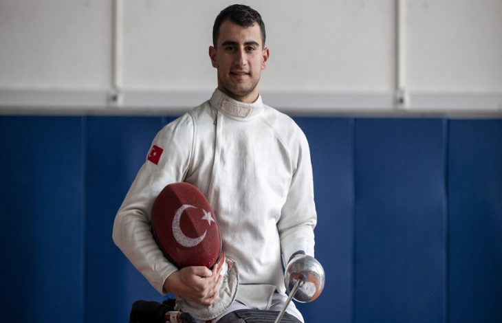 Türk bayrağı armalı beyaz eskrim kıyafeti giyen Hakan Akkaya, bir elinde ay-yıldızlı kılıfı, diğer elinde eskrim kılıcıyla spor salonunda poz veriyor.