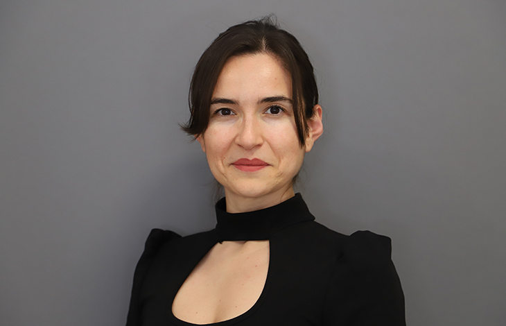 Görselde, sade gri bir arka plan önünde profesyonel bir portre çekimi yapılmış. Elif Türel, koyu renkli, yüksek yakalı ve göğüs kısmında küçük bir kesim detayı bulunan şık bir üst giyiyor. Saçları düzgünce geriye taranmış ve yüz hatlarını net şekilde ortaya çıkaran yumuşak bir ışık kullanılmış. Duruşu dik, ifadesi kendinden emin ve akademik bir profesyonelliği yansıtıyor.
