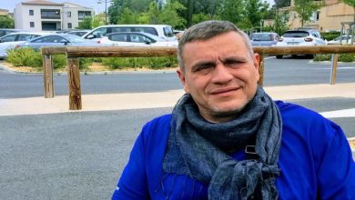 Ortopedik Engelli Astrolog Oğuzhan Ceyhan, üzerinde parlak mavi, kısa kollu bir tişört bulunuyor. Boynuna koyu gri, püsküllü bir atkı dolamış ve göğsünün önünde siyah askılı, açık gri renkli küçük bir omuz çantası taşıyor