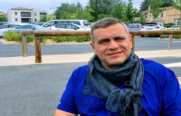 Ortopedik Engelli Astrolog Oğuzhan Ceyhan, üzerinde parlak mavi, kısa kollu bir tişört bulunuyor. Boynuna koyu gri, püsküllü bir atkı dolamış ve göğsünün önünde siyah askılı, açık gri renkli küçük bir omuz çantası taşıyor