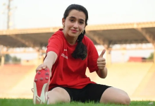 Görselin üst bölümünde, açık hava atletizm pistinde yeşil çim zemin üzerinde oturarak esneme hareketi yapan genç bir kadın sporcu yer almaktadır. Kırmızı tişört ve siyah şort giyen sporcu, bir eliyle ayağının ucundan tutarak esnerken diğer eliyle kameraya doğru baş parmağıyla onay (beğeni) işareti yapmakta ve gülümsemektedir. Arka planda stadyum tribünleri ve çatı yapısı görülmektedir.