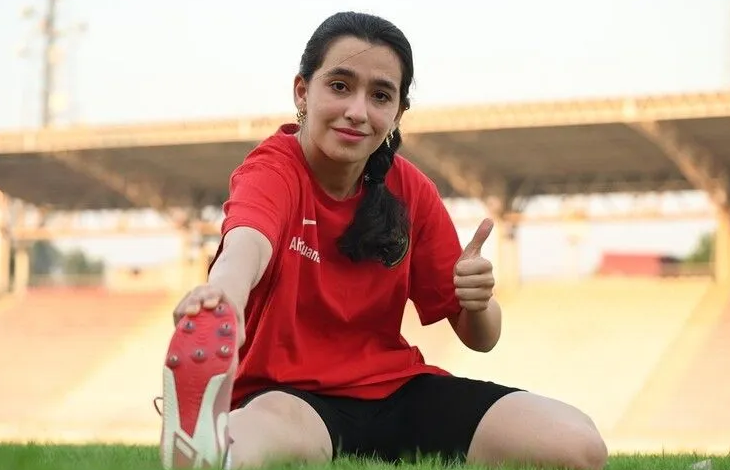 Görselin üst bölümünde, açık hava atletizm pistinde yeşil çim zemin üzerinde oturarak esneme hareketi yapan genç bir kadın sporcu yer almaktadır. Kırmızı tişört ve siyah şort giyen sporcu, bir eliyle ayağının ucundan tutarak esnerken diğer eliyle kameraya doğru baş parmağıyla onay (beğeni) işareti yapmakta ve gülümsemektedir. Arka planda stadyum tribünleri ve çatı yapısı görülmektedir.