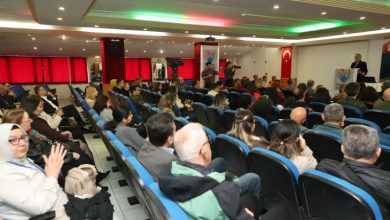 Katılımcılar seyirci koltuğunda kürsüde konuşan kadını dinliyor.