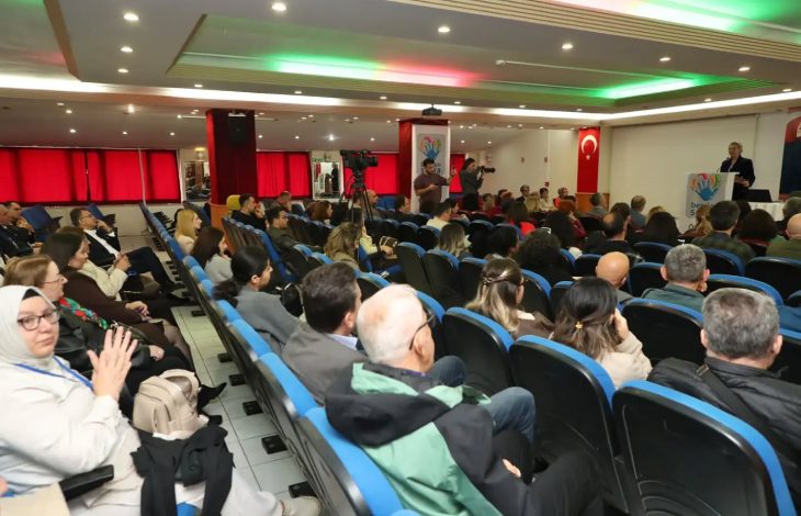 Katılımcılar seyirci koltuğunda kürsüde konuşan kadını dinliyor.