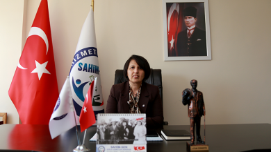 Masanın arkasında oturan SAHİM‑SEN Genel Başkanı Özlem Akarken, resmi bir ofis ortamında görülüyor. Masanın üzerinde Türk bayrağı ve SAHİM‑SEN bayrağı bulunan küçük masa flamaları, “Atam İzindeyiz” yazılı bir Atatürk büstü, bir takvim ve çalışma malzemeleri yer alıyor. Arka planda büyük bir Türk bayrağı, SAHİM‑SEN bayrağı ve duvarda Mustafa Kemal Atatürk portresi bulunuyor. Akarken, kurumunu temsil eden sakin ve profesyonel bir duruşla kameraya bakıyor.