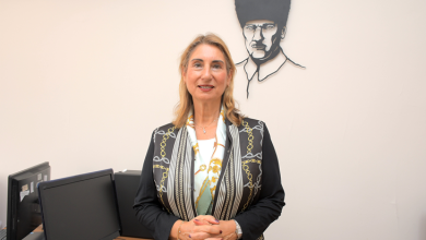 Kapalı bir mekânda, muhtemelen bir bilgisayar laboratuvarı ya da eğitim odasında duran Prof. Dr. Gülser Kılınç, kameraya doğru sakin ve kendinden emin bir ifadeyle bakıyor. Üzerinde siyah zeminli, altın ve beyaz zincir motifleriyle süslenmiş şık bir ceket bulunuyor. Boynunda açık renkli bir şal var; bu şal görünümüne zarif bir dokunuş katıyor. Arka planda, duvarda Mustafa Kemal Atatürk’ün siyah hat çizimiyle yapılmış portresi dikkat çekiyor. Çevrede masa üstü bilgisayarlar, klavyeler ve monitörler yer alıyor; bu da ortamın akademik veya kurumsal bir eğitim alanı olduğunu düşündürüyor. Prof. Dr. Gülser Kılınç fotoğrafta hem profesyonel hem de sıcak bir duruş sergiliyor; arka plandaki Atatürk portresi ise mekâna anlamlı bir kültürel vurgu katıyor.