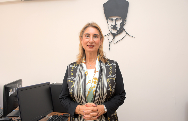 Kapalı bir mekânda, muhtemelen bir bilgisayar laboratuvarı ya da eğitim odasında duran Prof. Dr. Gülser Kılınç, kameraya doğru sakin ve kendinden emin bir ifadeyle bakıyor. Üzerinde siyah zeminli, altın ve beyaz zincir motifleriyle süslenmiş şık bir ceket bulunuyor. Boynunda açık renkli bir şal var; bu şal görünümüne zarif bir dokunuş katıyor. Arka planda, duvarda Mustafa Kemal Atatürk’ün siyah hat çizimiyle yapılmış portresi dikkat çekiyor. Çevrede masa üstü bilgisayarlar, klavyeler ve monitörler yer alıyor; bu da ortamın akademik veya kurumsal bir eğitim alanı olduğunu düşündürüyor. Prof. Dr. Gülser Kılınç fotoğrafta hem profesyonel hem de sıcak bir duruş sergiliyor; arka plandaki Atatürk portresi ise mekâna anlamlı bir kültürel vurgu katıyor.