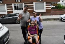 Görselin üst bölümünde, sokakta özel destekli çocuk tekerlekli sandalyesinde oturan serebral palsili küçük kız çocuğu Yağmur ve hemen arkasında duran anne ile babası yer alıyor. Sandalyede oturan Yağmur, fuşya renkli tişörtü ve bej şortuyla hafifçe gülümseyerek sol elini havaya kaldırıp selam veriyor. Arkasındaki gri polo yaka tişörtlü babası ile açık mor tişörtlü annesi de neşeyle birer ellerini kaldırarak kameraya el sallıyor. Arka planda modern bir apartman binası, sokakta park halinde duran gri ve beyaz renkli otomobiller görülüyor.