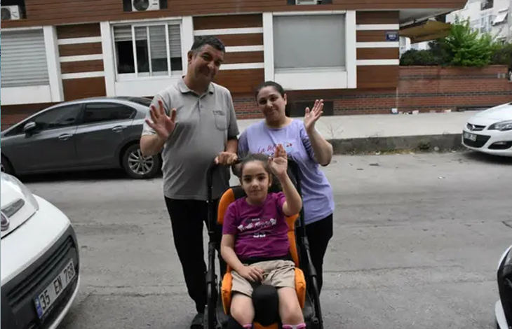 Görselin üst bölümünde, sokakta özel destekli çocuk tekerlekli sandalyesinde oturan serebral palsili küçük kız çocuğu Yağmur ve hemen arkasında duran anne ile babası yer alıyor. Sandalyede oturan Yağmur, fuşya renkli tişörtü ve bej şortuyla hafifçe gülümseyerek sol elini havaya kaldırıp selam veriyor. Arkasındaki gri polo yaka tişörtlü babası ile açık mor tişörtlü annesi de neşeyle birer ellerini kaldırarak kameraya el sallıyor. Arka planda modern bir apartman binası, sokakta park halinde duran gri ve beyaz renkli otomobiller görülüyor.