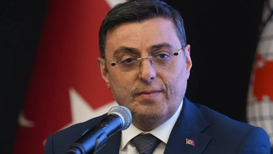 Koyu renk takım elbise giyen, gözlüklü Serkan Bayram mikrofonla resmi bir açıklama yapıyor. Arkasında Türk bayrağı bulunuyor. Ortam, ciddi ve kurumsal bir basın toplantısı ya da resmi etkinlik havası taşıyor.