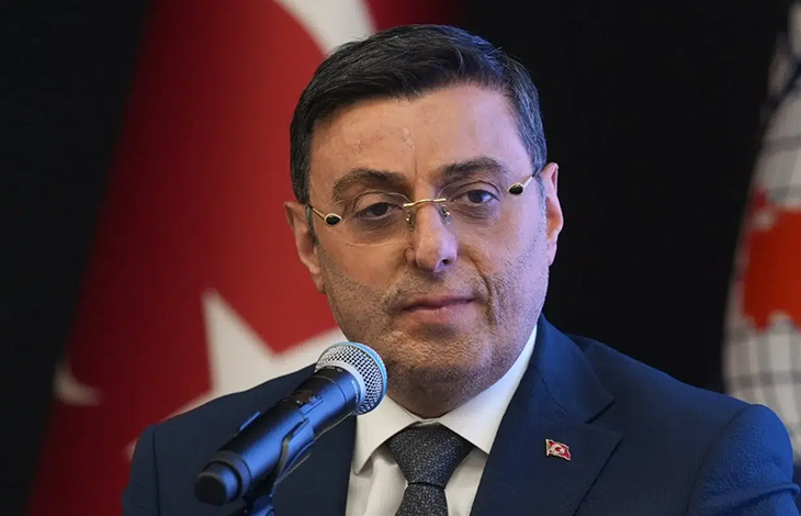 Koyu renk takım elbise giyen, gözlüklü Serkan Bayram mikrofonla resmi bir açıklama yapıyor. Arkasında Türk bayrağı bulunuyor. Ortam, ciddi ve kurumsal bir basın toplantısı ya da resmi etkinlik havası taşıyor.