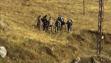 Görselin üst bölümünde, sarı otlarla kaplı engebeli ve açık bir arazide yürüyen bir grup insan ve aralarında askerî kamuflajlı personelin bulunduğu bir kalabalık yer almaktadır. Sağ tarafta metal bir elektrik direği ve arazide patika benzeri yollar dikkat çekmektedir.