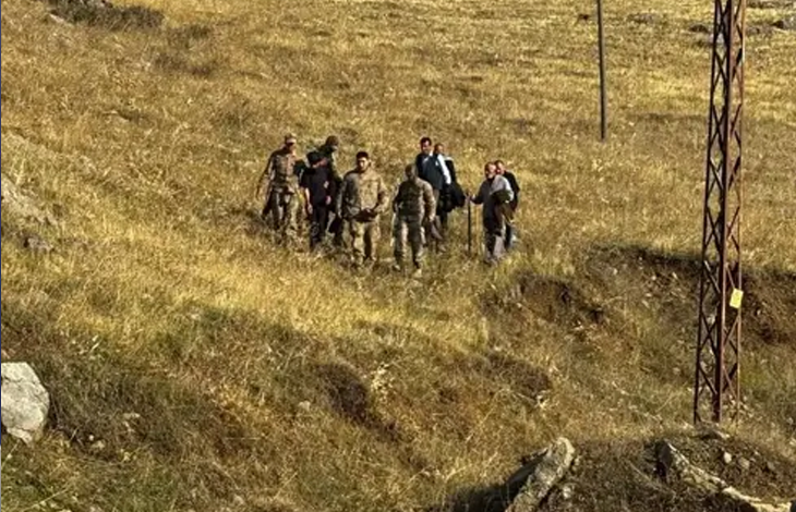 Görselin üst bölümünde, sarı otlarla kaplı engebeli ve açık bir arazide yürüyen bir grup insan ve aralarında askerî kamuflajlı personelin bulunduğu bir kalabalık yer almaktadır. Sağ tarafta metal bir elektrik direği ve arazide patika benzeri yollar dikkat çekmektedir.