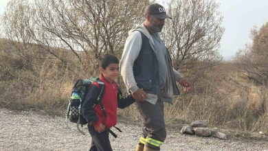 Görselde, kırsal veya dağlık bir yolda el ele tutuşarak yürüyen sırt çantalı bir çocuk ile yetişkin bir erkek yer almaktadır.