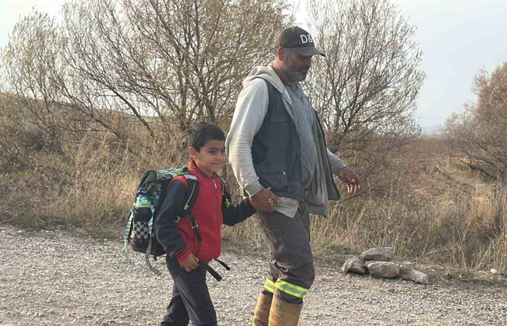 Görselde, kırsal veya dağlık bir yolda el ele tutuşarak yürüyen sırt çantalı bir çocuk ile yetişkin bir erkek yer almaktadır.
