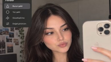 Bir genç kadın, elinde akıllı telefonuyla selfie çekerken burun şekli, yüz şekli, güzelleştirme ve doğal görünüm gibi sosyal medya filtrelerini gösteren bir arayüz eşliğinde yüz hatlarını inceliyor.